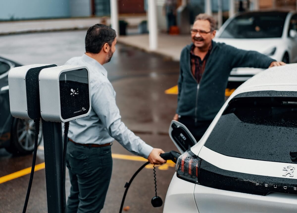A driver plugging an electric car into workplace chargers while talking to a colleague